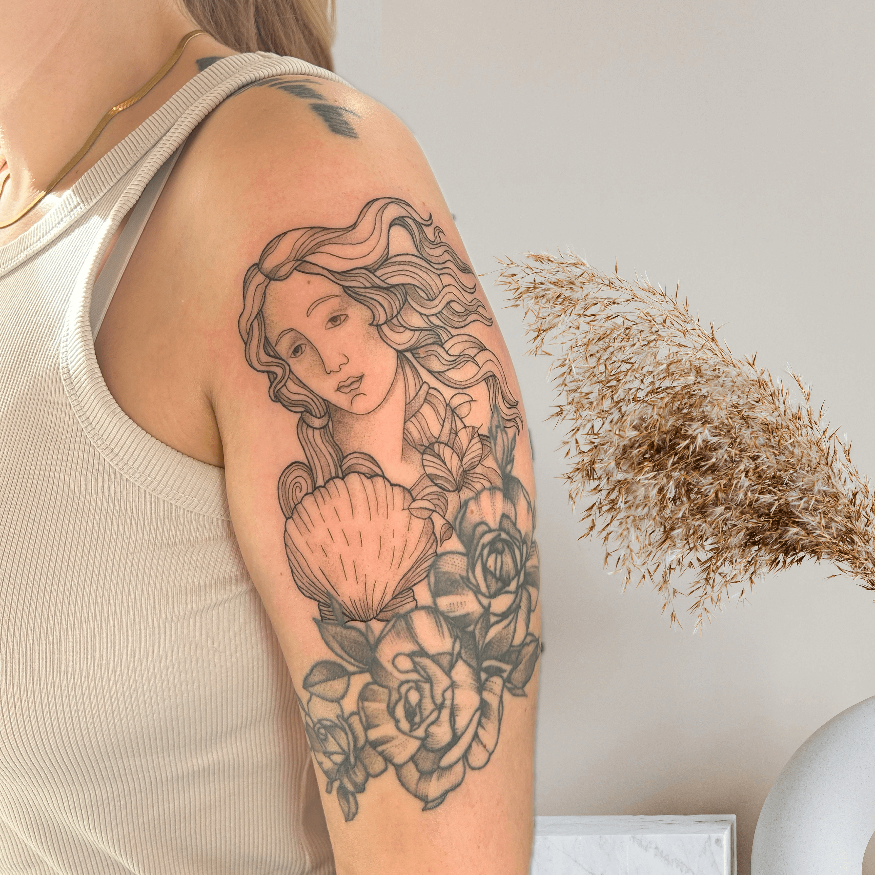 Tattoo einer weiblichen Figur mit langen Haaren, die eine Muschel hält, umgeben von Rosen auf dem Oberarm.