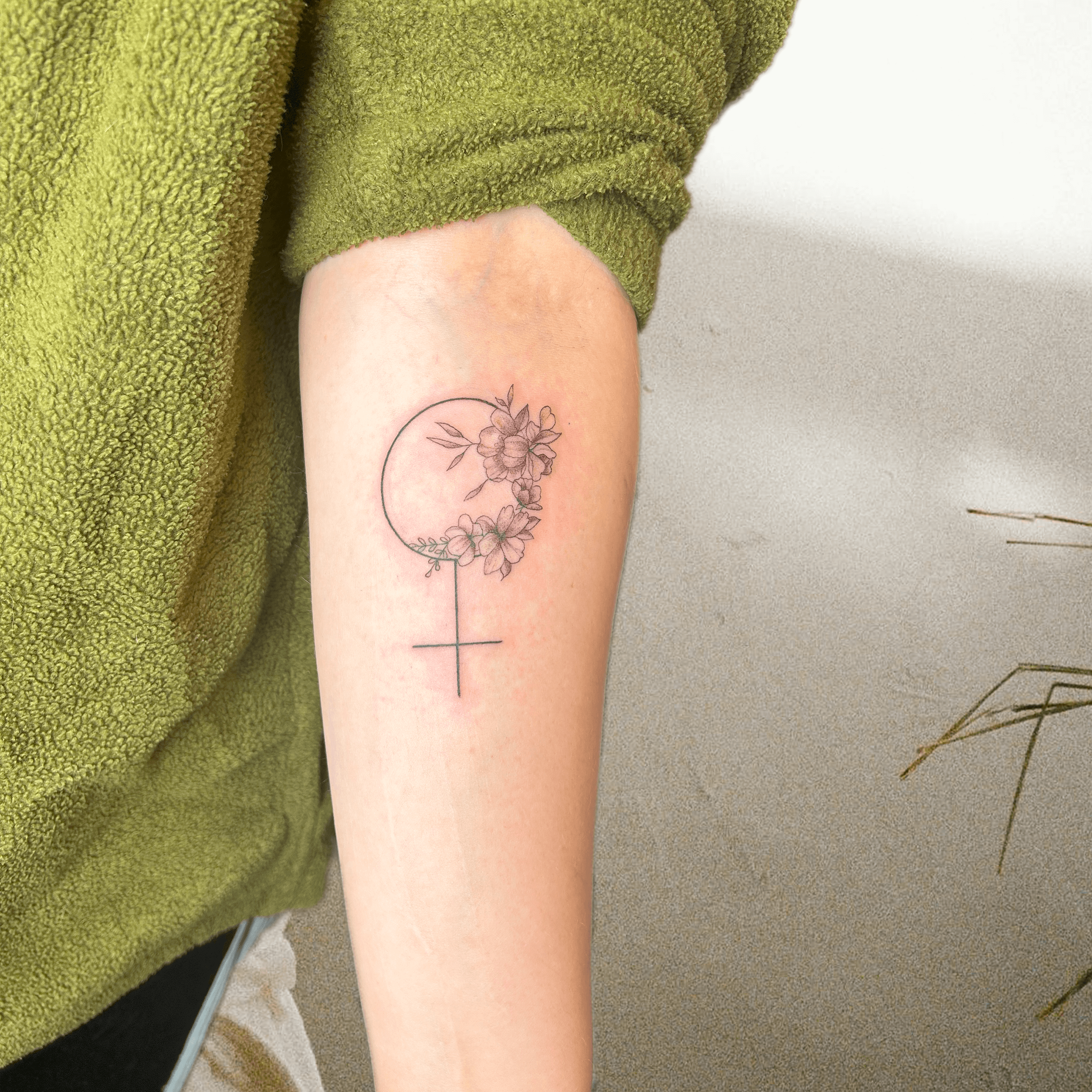 Tattoo eines femininen Symbols mit Blumenverzierung auf dem Unterarm.