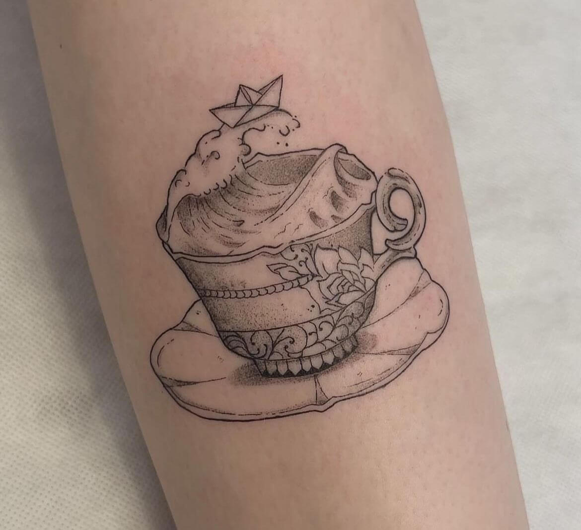 Tattoo einer dekorativen Vintage-Tasse, die von einer Welle überflutet wird, mit einem Origami-Papierboot darauf.