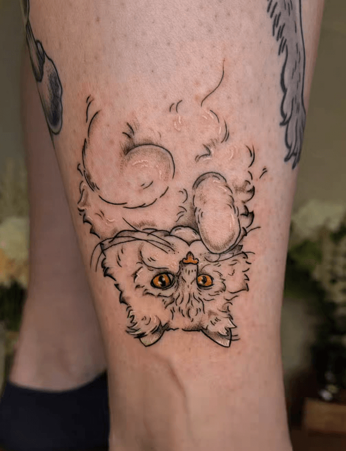 Tattoo einer flauschigen Katze mit gelben Augen, die auf dem Rücken liegt und verspielt in die Kamera schaut – feine Linien, Detailzeichnung auf der Haut